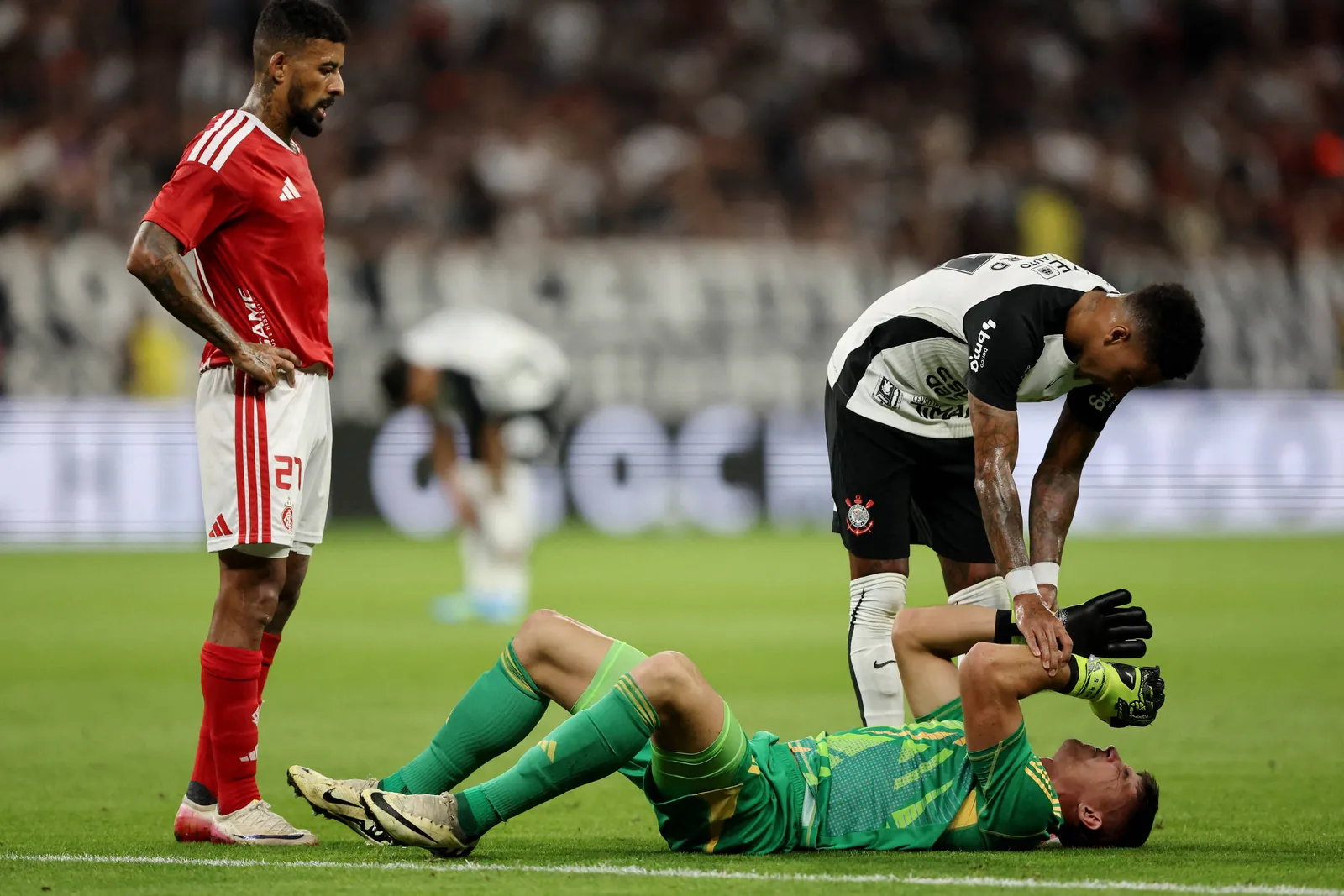 Jogador de futebol vestido com uniforme verde está deitado no gramado segurando a cabeça, enquanto companheiro de time com uniforme preto e branco se inclina para ajudá-lo. Jogador adversário com uniforme vermelho observa a cena em pé.
