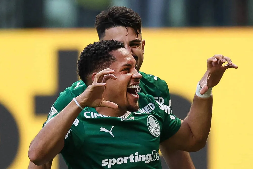 Dois jogadores do Palmeiras comemoram um gol. O jogador Ã frente sorri e levanta os braÃ§os, enquanto o jogador atrÃ¡s o abraÃ§a. Ambos vestem uniforme verde com patrocÃ­nios visÃ­veis.