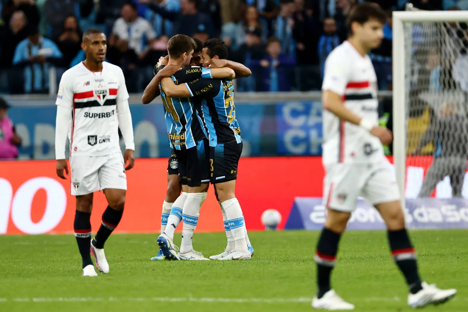 Dois jogadores do Grêmio abraçam-se no centro do campo celebrando gol, enquanto jogadores do São Paulo caminham cabisbaixos próximos à área de gol. Torcida e placas publicitárias aparecem ao fundo.